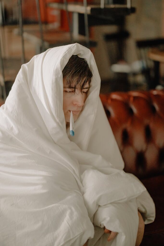 Person bundled in a blanket with a thermometer, indoors, feeling unwell.
